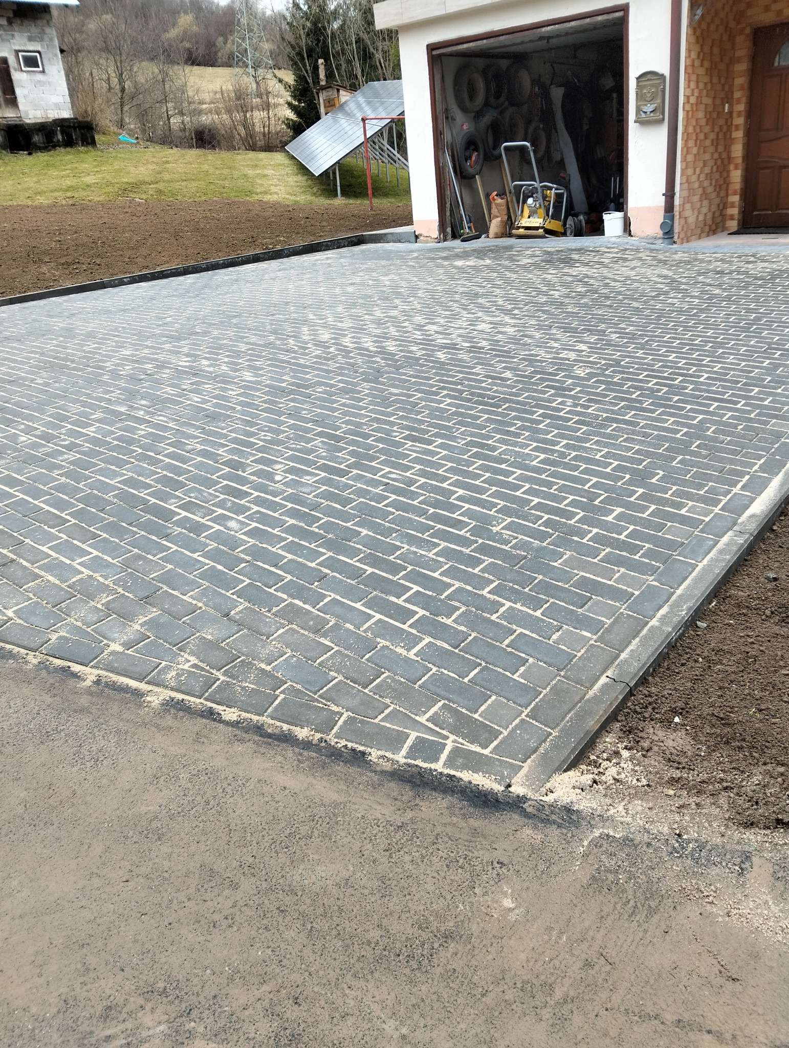 Nowo ułożona, szara kostka brukowa w jodełkę na podjeździe, z widocznym garażem i przygotowanym terenem pod trawnik. Kostka wysypana piaskiem.