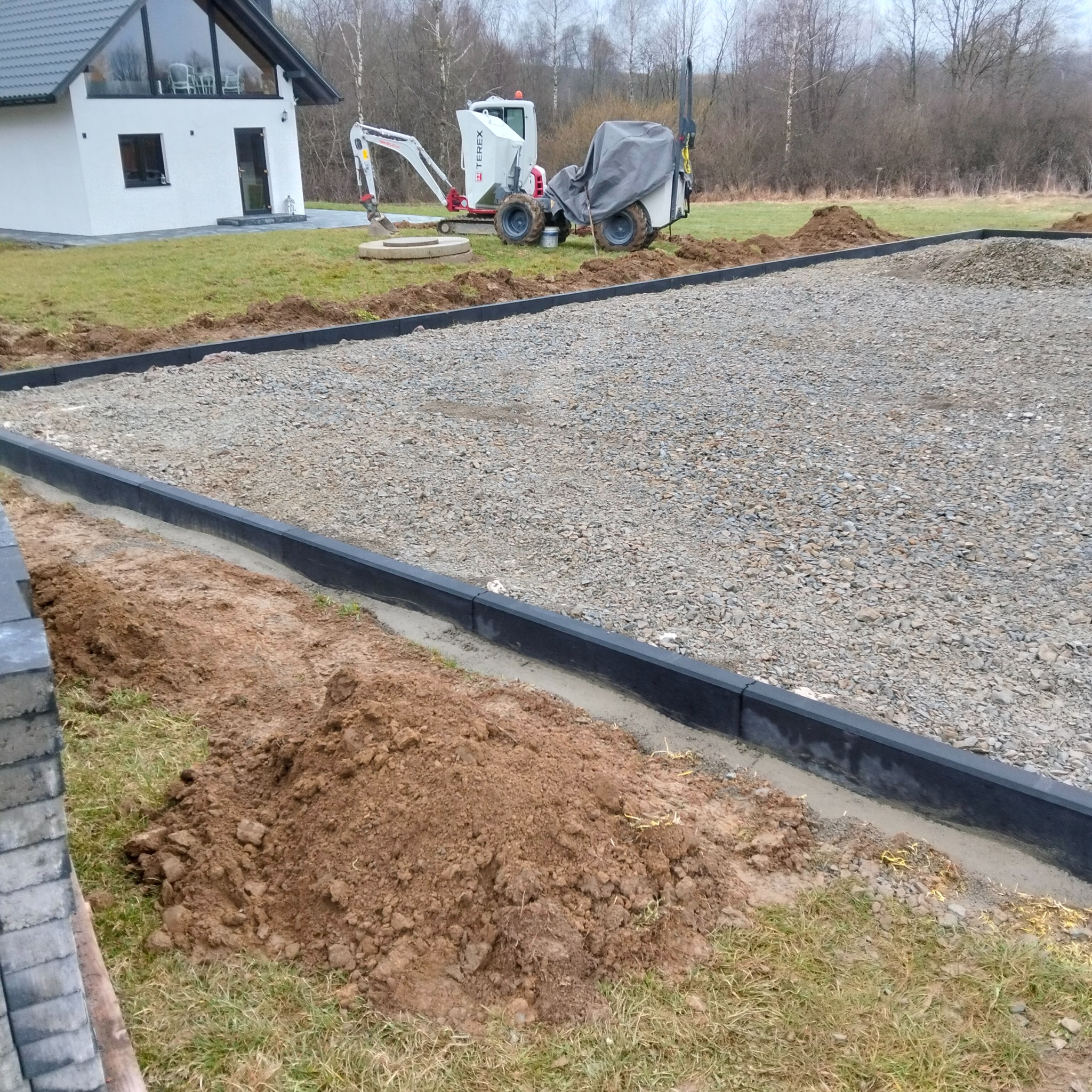 Przygotowany teren pod kostkę brukową: podsypka z kamienia, obrzeża z betonu. W tle dom i maszyny budowlane. Etap prac przed ułożeniem kostki.