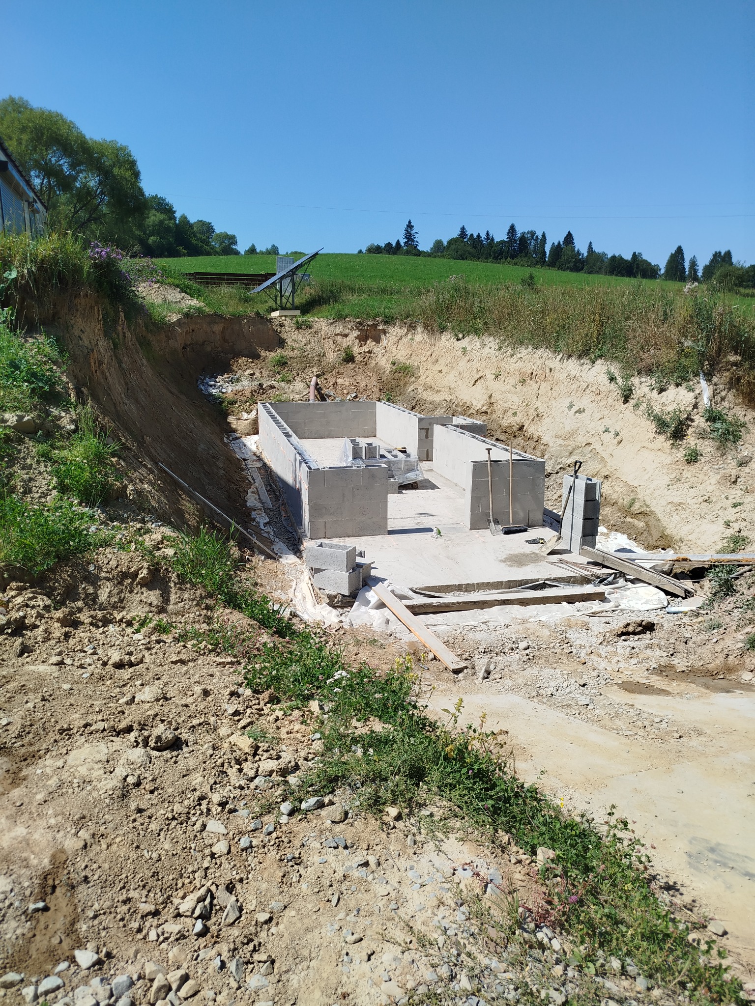 Fundamenty domu jednorodzinnego w wykopie, widok z góry, narzędzia budowlane obok. W tle zielone wzgórza i błękitne niebo.