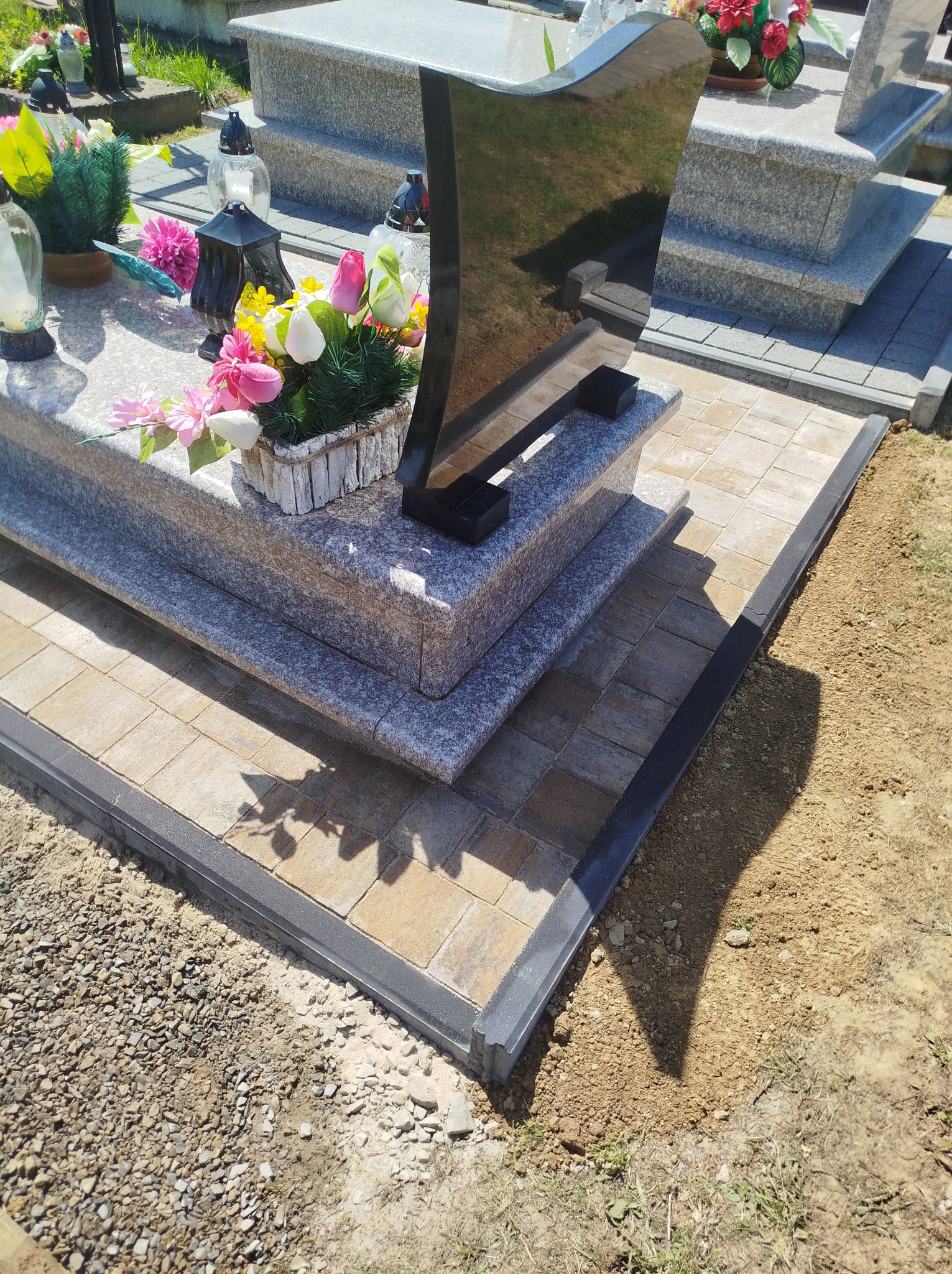 Ułożona kostka brukowa wokół nagrobka z kwiatami, wykończona czarnym obrzeżem. Widok na fragment cmentarza w słoneczny dzień. Prace brukarskie przy pomniku.