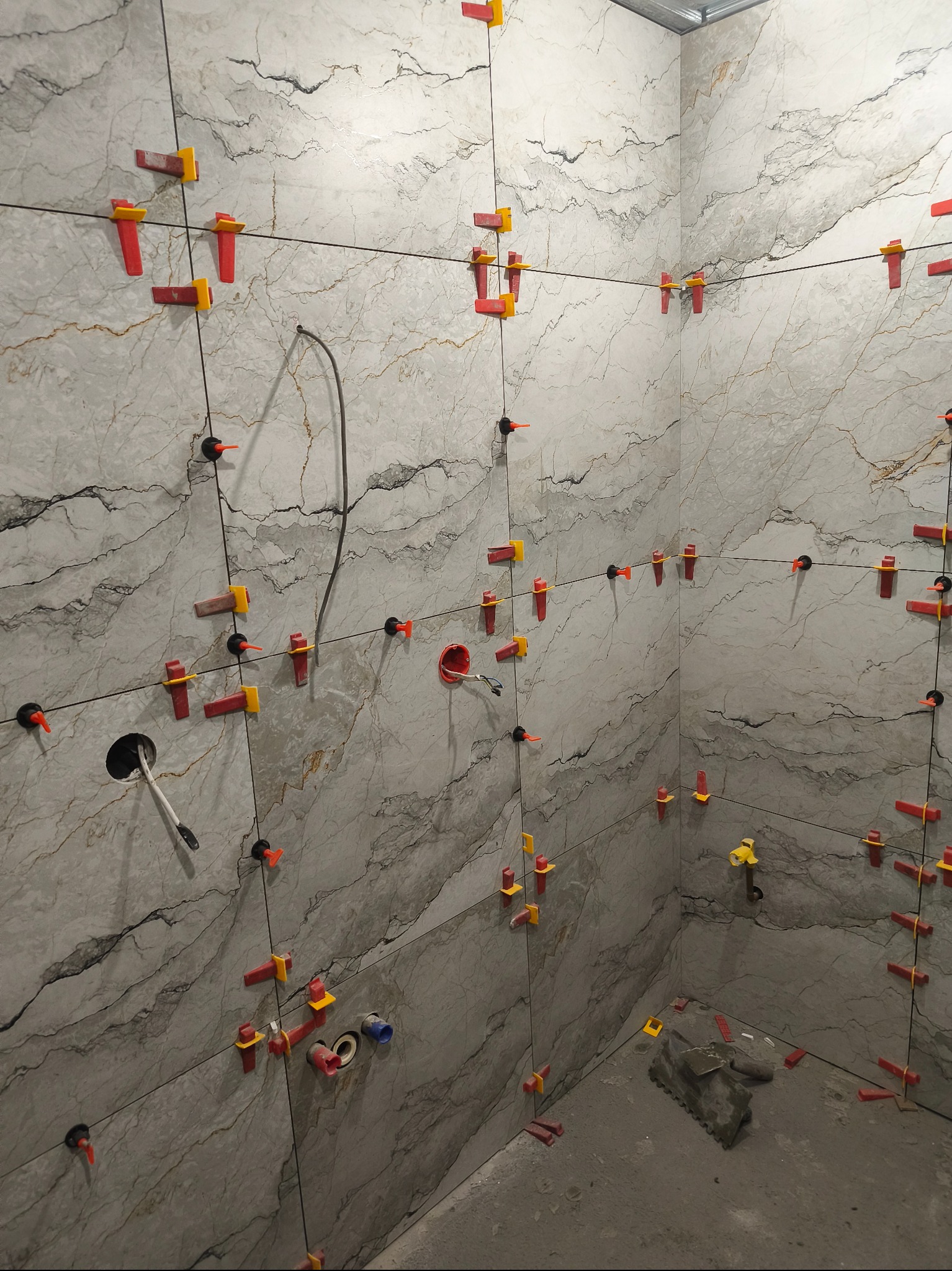 Ściany łazienki w trakcie układania płytek imitujących marmur, z użyciem systemu poziomowania płytek (klipsy i kliny). Widoczne otwory na instalacje i narzędzia glazurnicze.