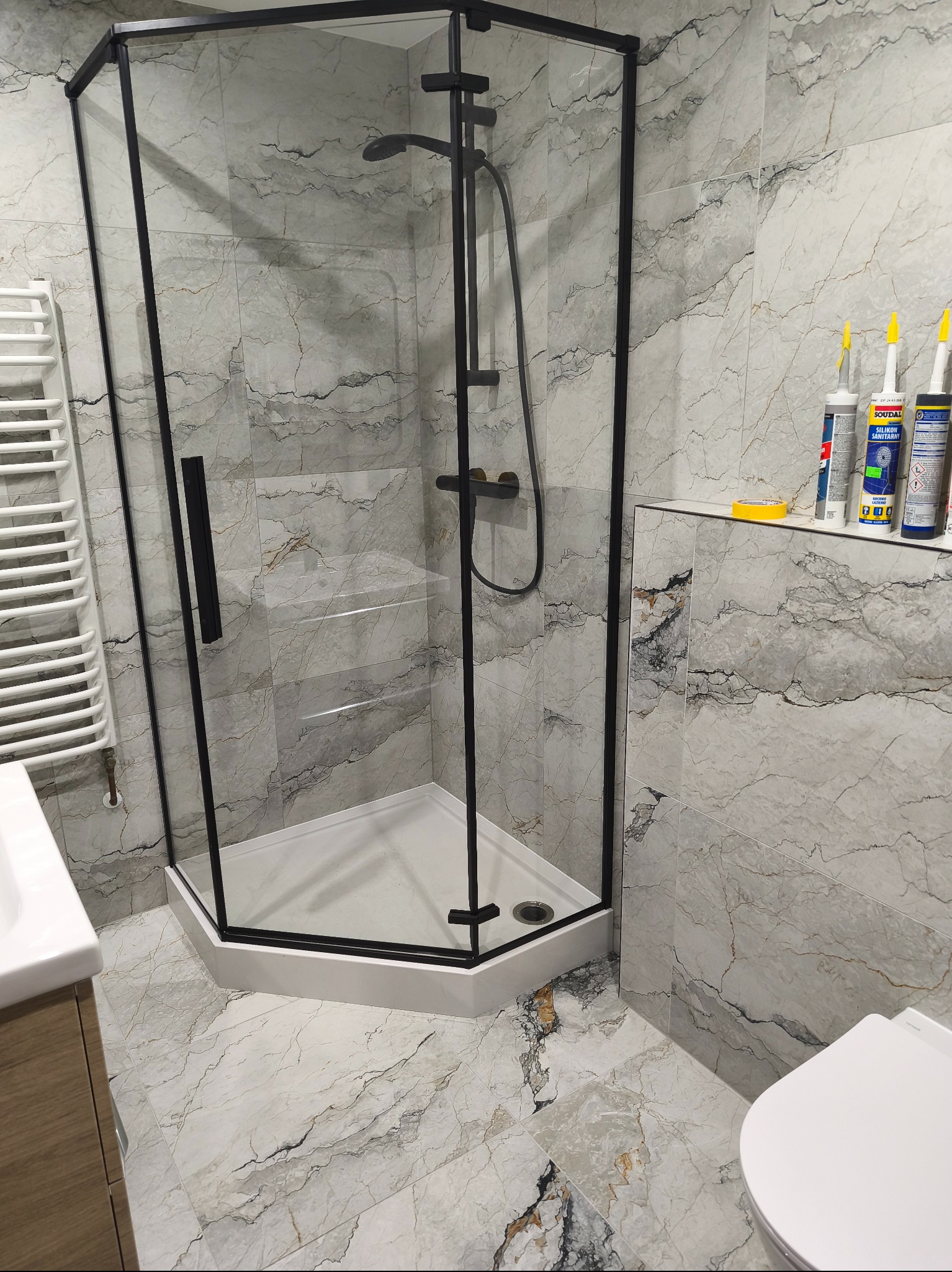 Nowoczesna kabina prysznicowa z czarnymi profilami na tle marmurowych płytek w łazience. Widoczne silikony sanitarne na półce.