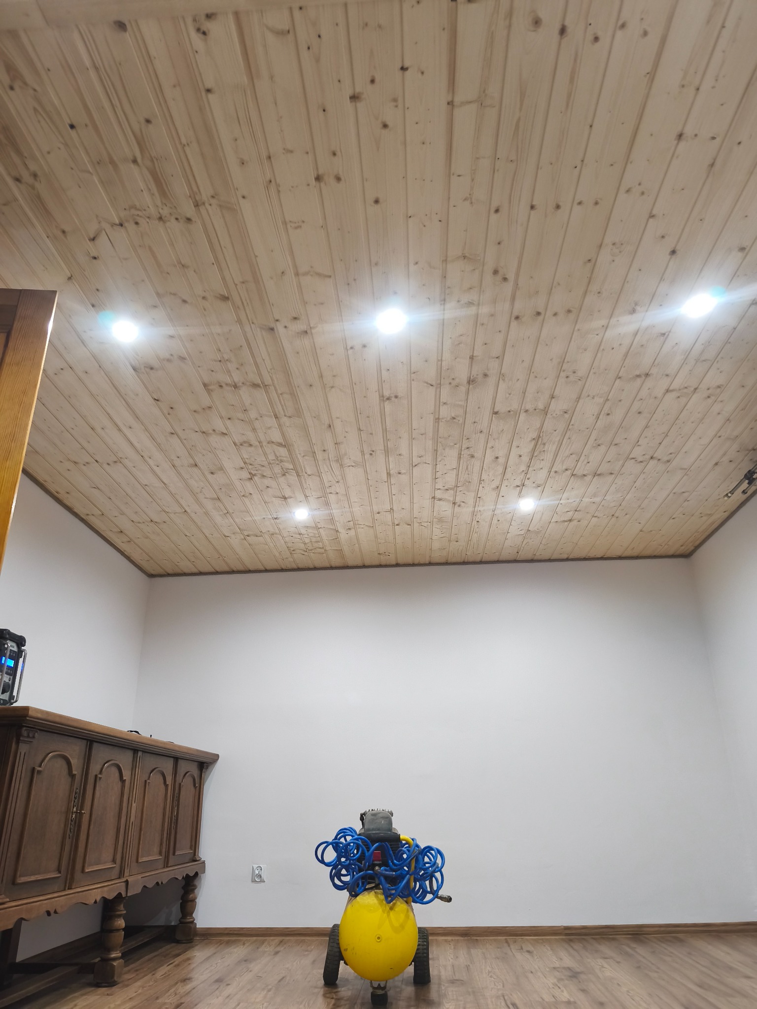 Drewniany sufit z wbudowanymi punktowymi lampami w jasnym pomieszczeniu. Na podłodze kompresor i drewniana komoda. Ujęcie z dołu, podkreślające wykończenie.