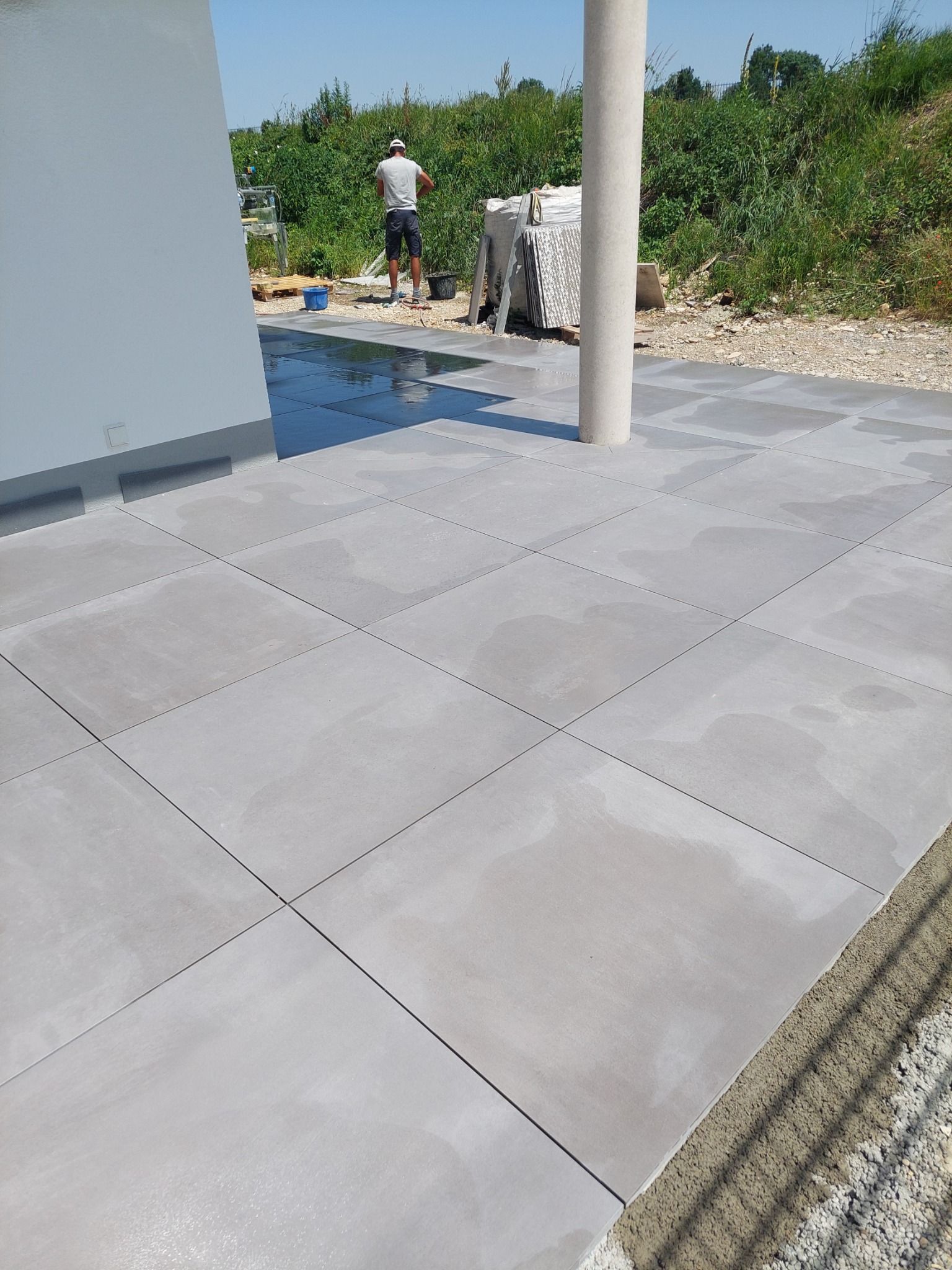 Świeżo ułożona, mokra, szara nawierzchnia z płyt betonowych na posesji, z widocznym słupem i pracownikiem w tle, w trakcie czyszczenia po ułożeniu.
