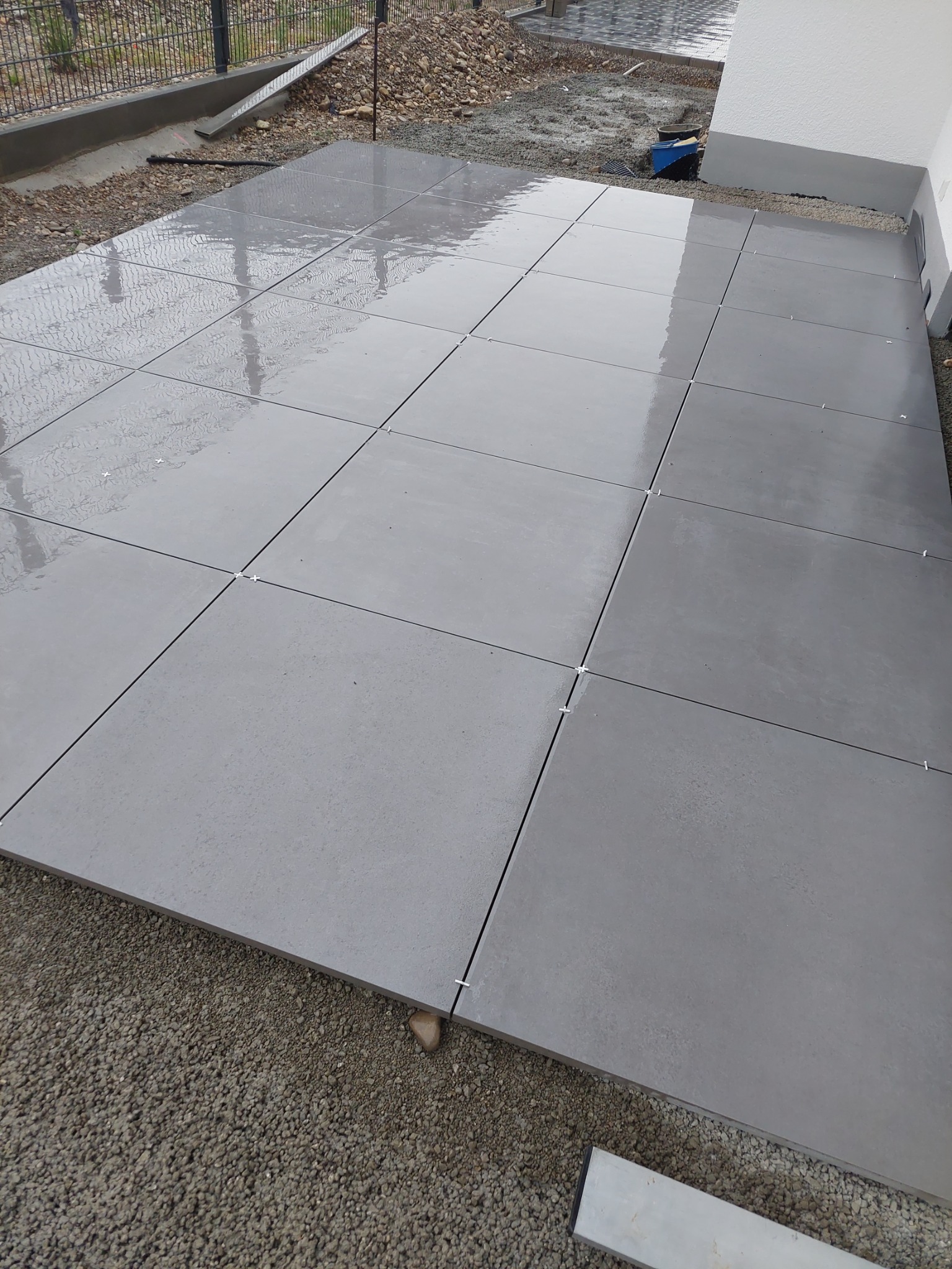 Świeżo ułożone, mokre, szare płyty tarasowe z krzyżykami dystansowymi, widoczne drobne kamienie pod spodem, obok ściana budynku.