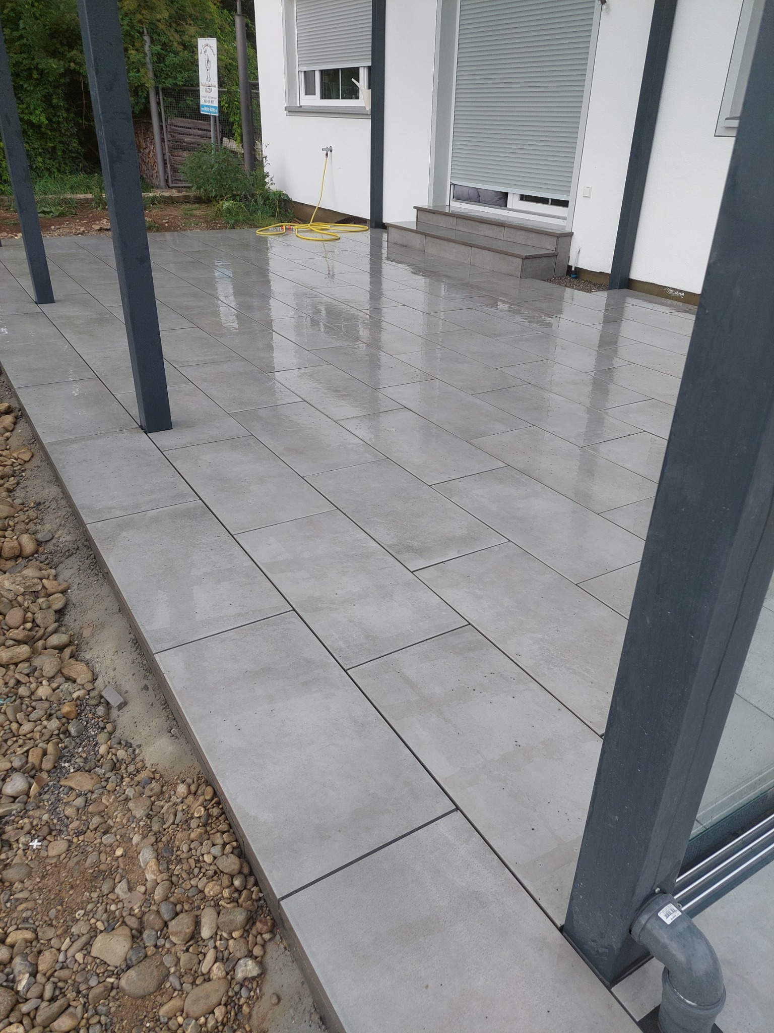 Świeżo ułożona, mokra kostka brukowa w odcieniach szarości na tarasie z widocznymi słupami konstrukcyjnymi z odprowadzeniem wody i fragmentem ogrodu w tle.