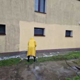 Pracownik w żółtym płaszczu przeciwdeszczowym czyści elewację domu myjką ciśnieniową, widoczne różnice w kolorze elewacji przed i po czyszczeniu, zacieki na trawie.