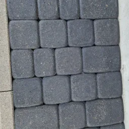 Ułożona, szara kostka brukowa o zaokrąglonych krawędziach, widoczna z bliska, z fragmentem jasnoszarego obrzeża po lewej stronie i białym elementem po prawej.