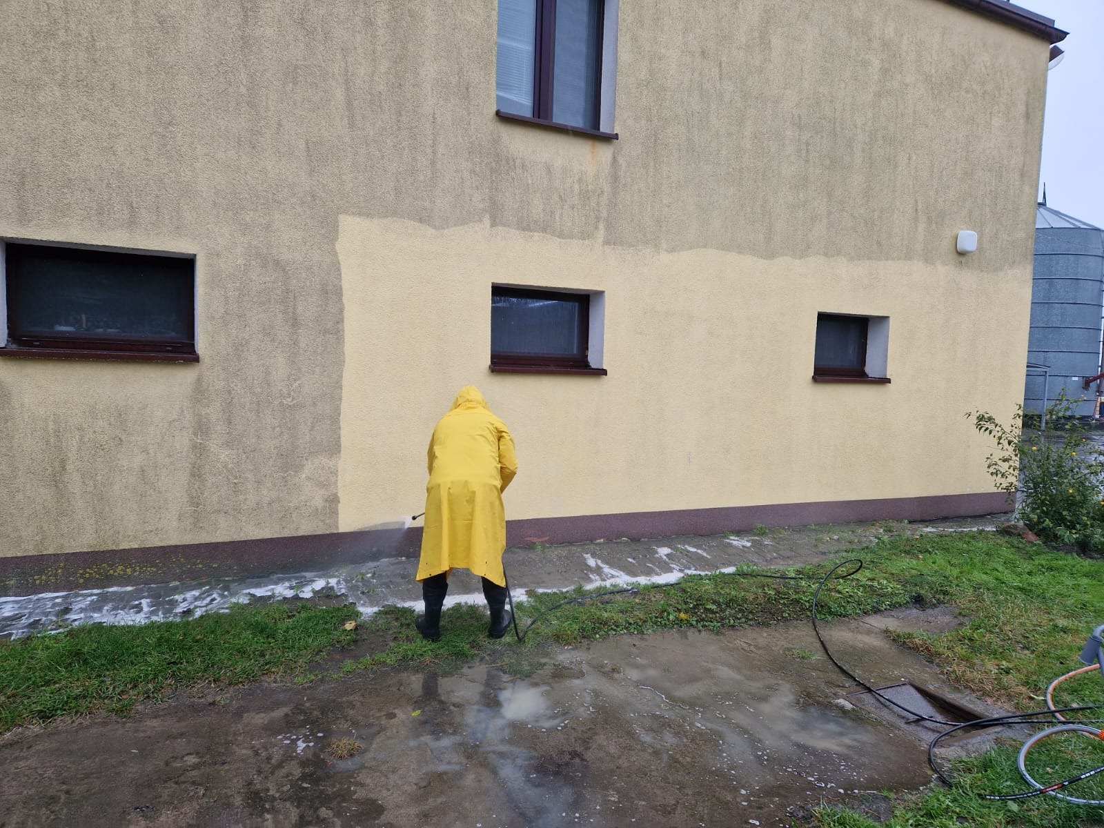 Pracownik w żółtym płaszczu przeciwdeszczowym czyści elewację domu myjką ciśnieniową, widoczne różnice w kolorze elewacji przed i po czyszczeniu, zacieki na trawie.