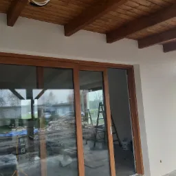 Nowe, drewniane okno przesuwne z widokiem na posesję w trakcie prac wykończeniowych, widoczna drabina i wiadro z farbą.