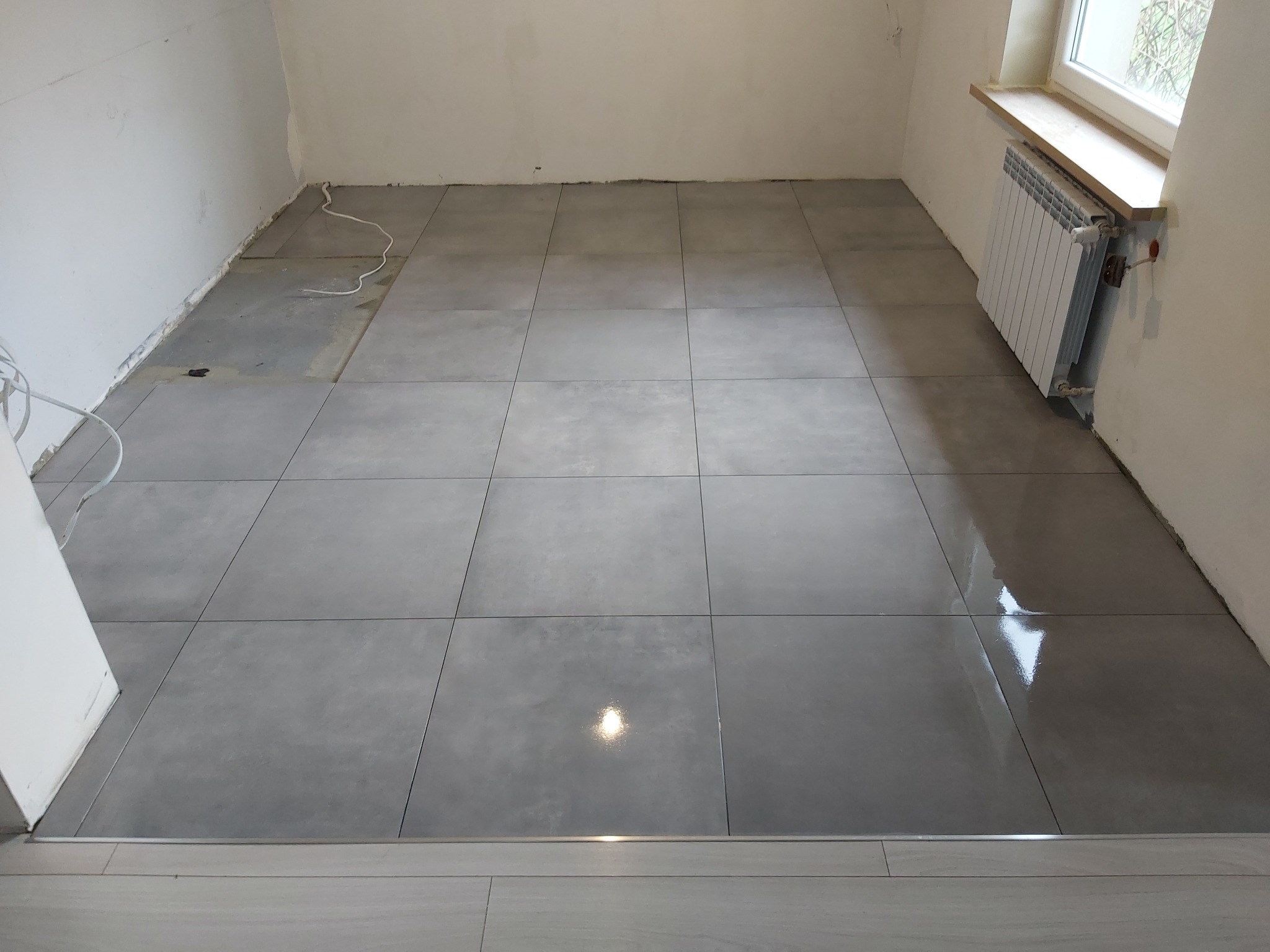 Ułożone, duże, szare płytki podłogowe w pomieszczeniu z grzejnikiem pod oknem, widoczne niedokończone prace wykończeniowe przy ścianach i listwach przypodłogowych.