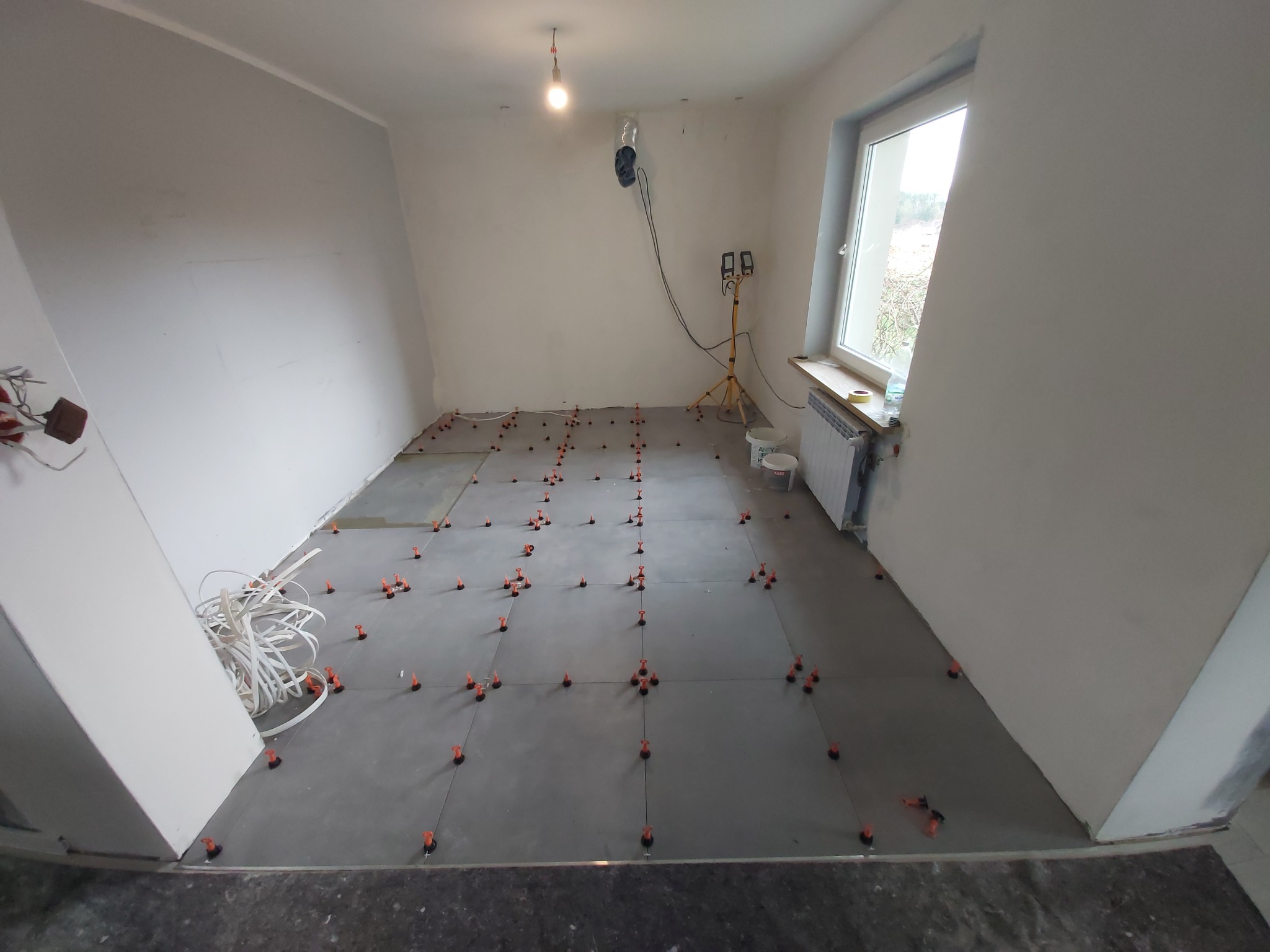 Układanie szarych płytek podłogowych w pomieszczeniu, z użyciem pomarańczowych klinów poziomujących; widoczna instalacja oświetleniowa i grzejnik pod oknem.