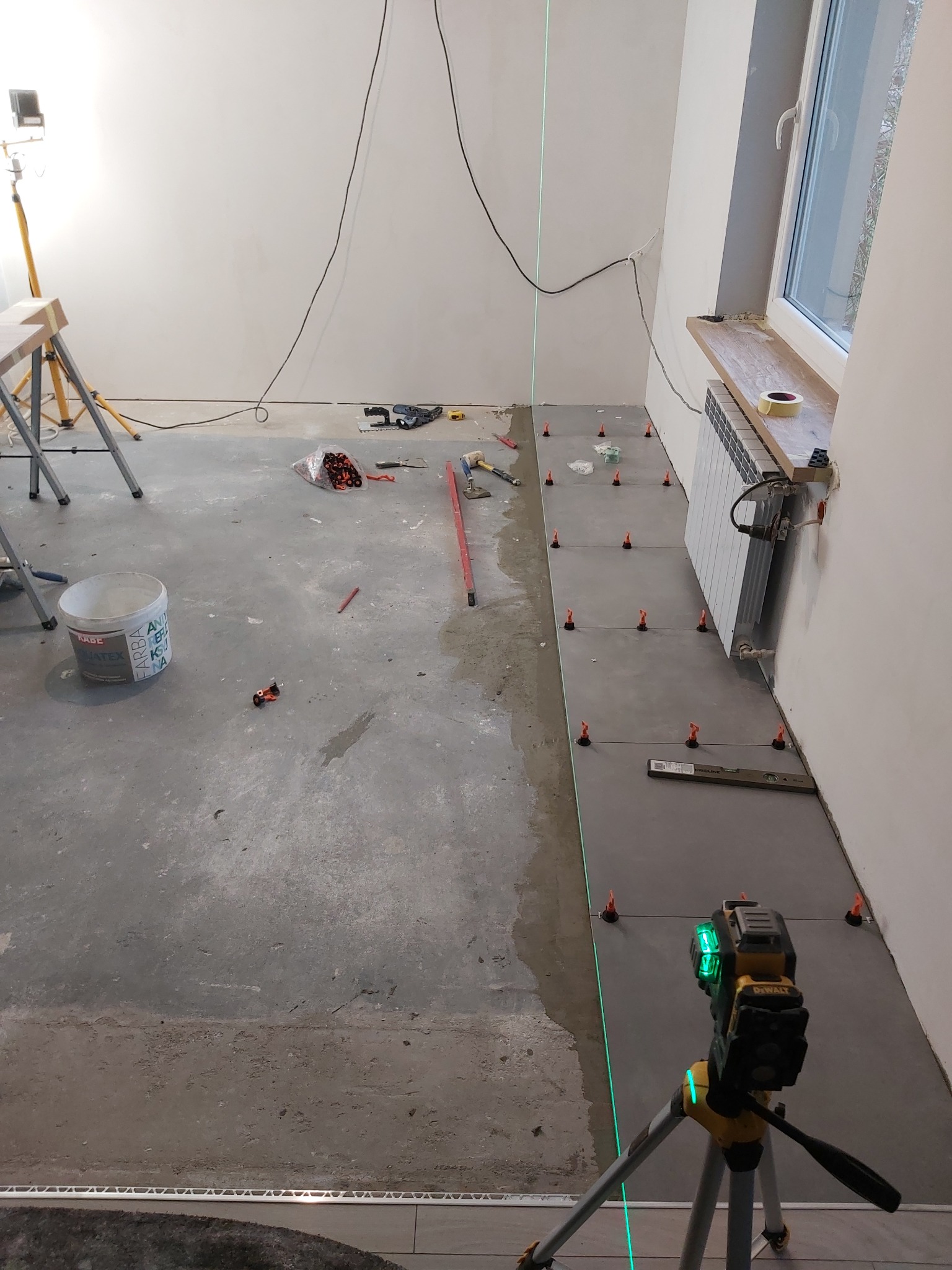 Układanie szarych płytek podłogowych w trakcie remontu, widoczny laser krzyżowy DeWalt, poziomica, wiadro z farbą Kabe, krzyżyki dystansowe i narzędzia glazurnicze.
