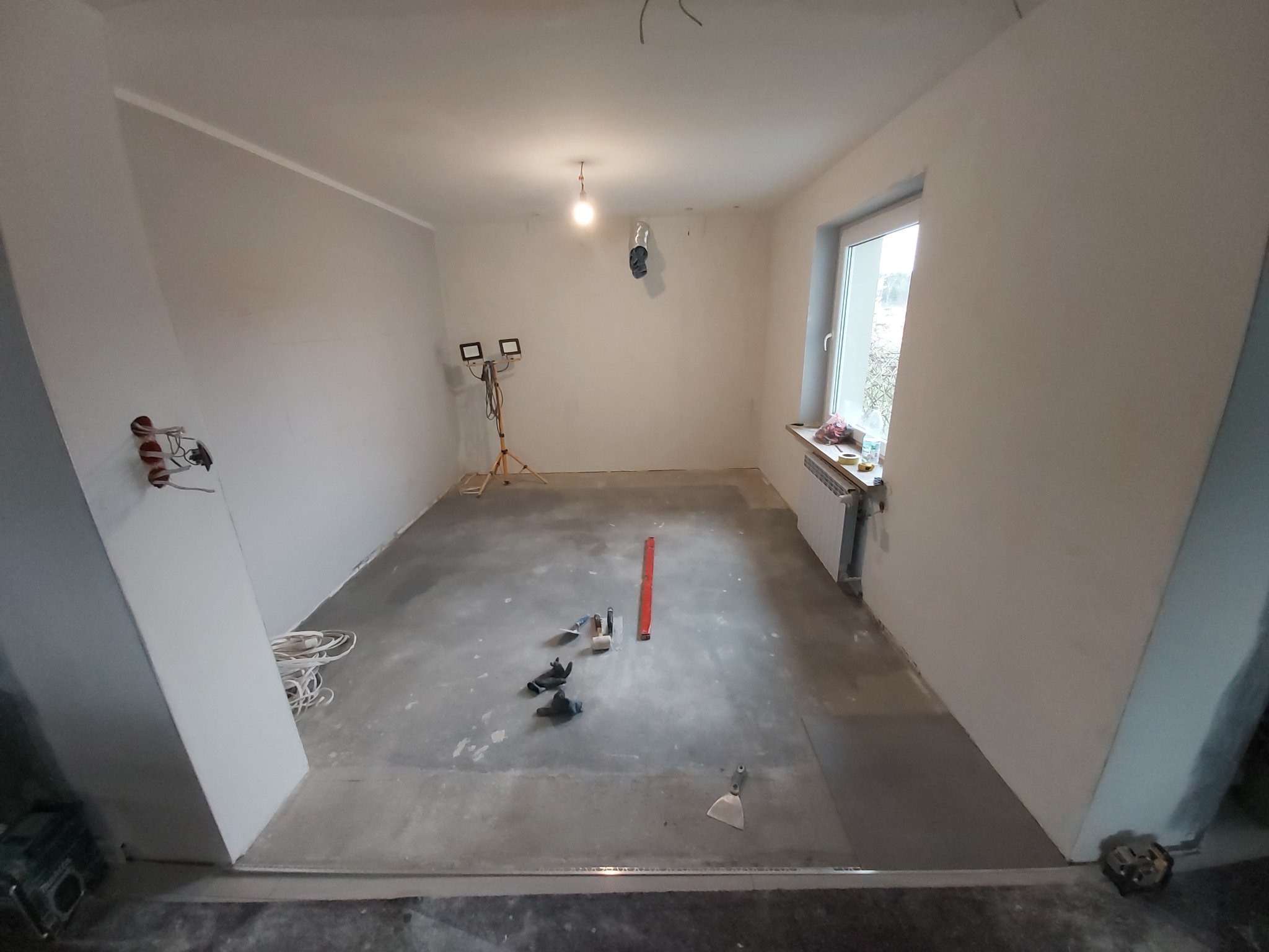 Przygotowane do wykończenia wnętrze pokoju z surowymi ścianami, szarym podkładem podłogowym, narzędziami na podłodze i lampą budowlaną na statywie