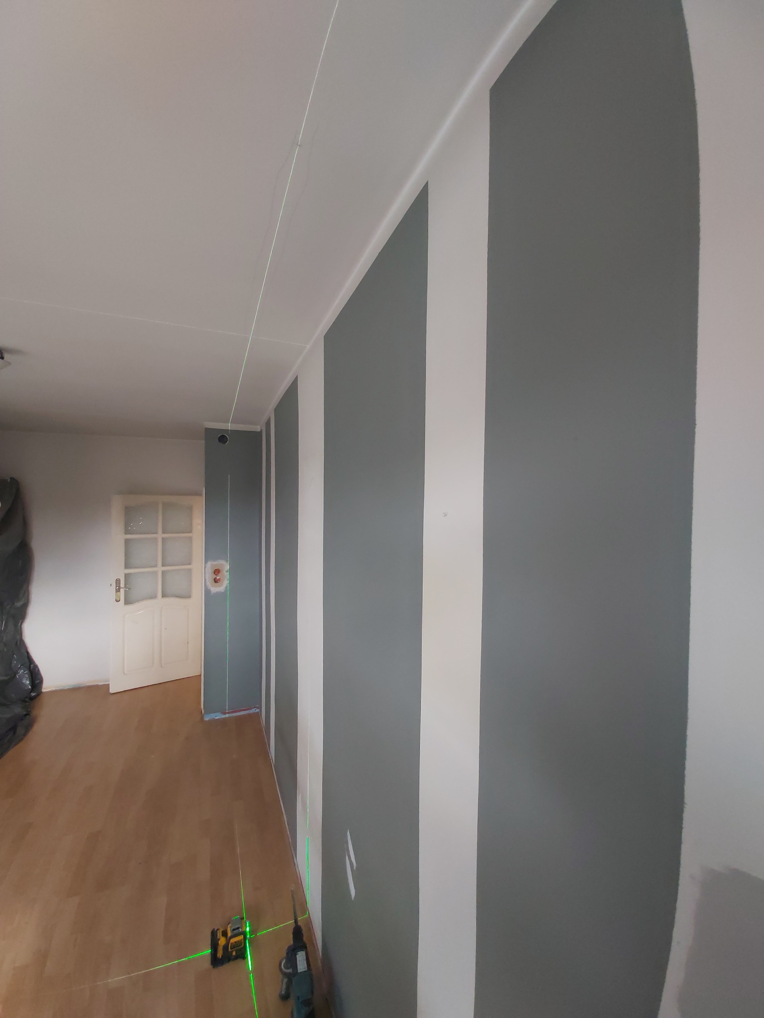 Świeżo pomalowana ściana w szaro-białe pasy, z widocznym laserowym niwelatorem wyznaczającym pion, drzwi w tle.