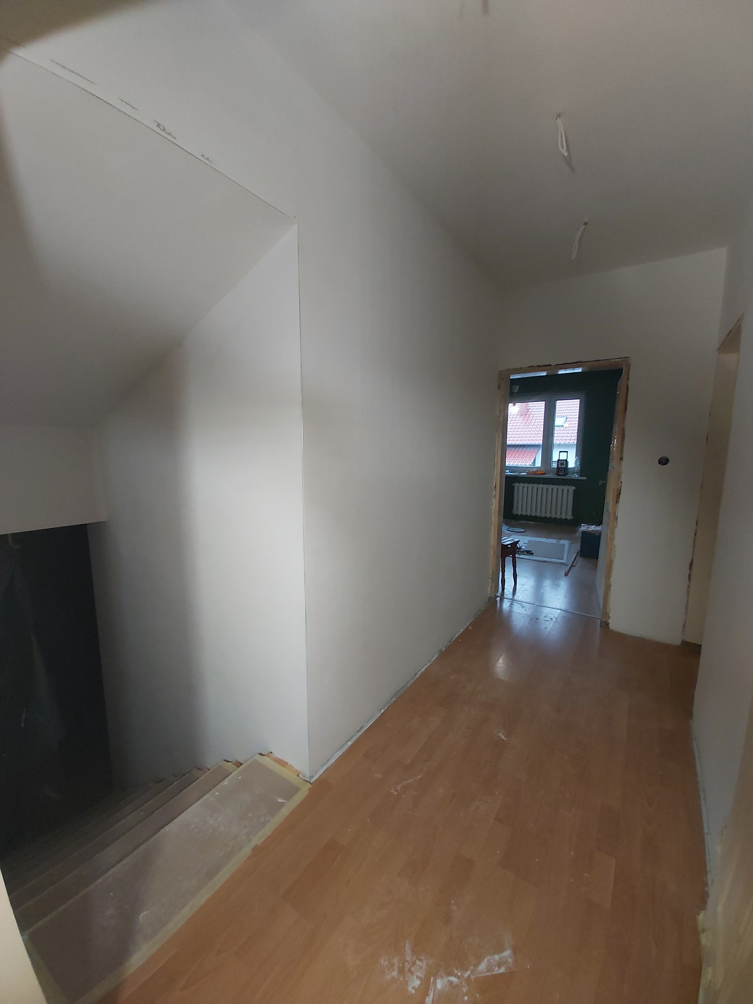 Korytarz w trakcie prac wykończeniowych: świeżo pomalowane ściany na biało, drewniana podłoga, schody w lewo, widoczne kable wystające z sufitu, drzwi prowadzące do innego pokoju.