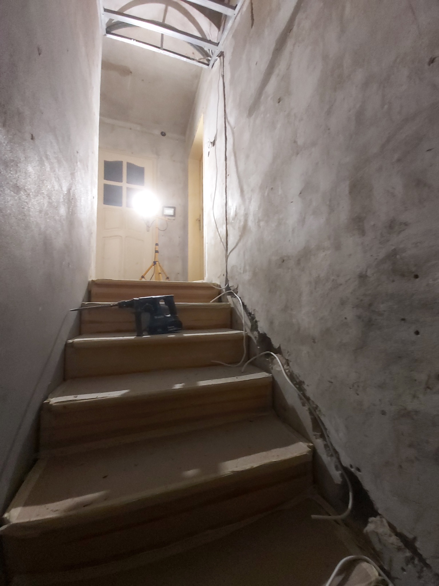 Wnętrze klatki schodowej w trakcie remontu, widoczne surowe ściany z ubytkami tynku, drewniane schody oraz wiertarka udarowa leżąca na jednym ze stopni, w tle lampa budowlana na statywie.
