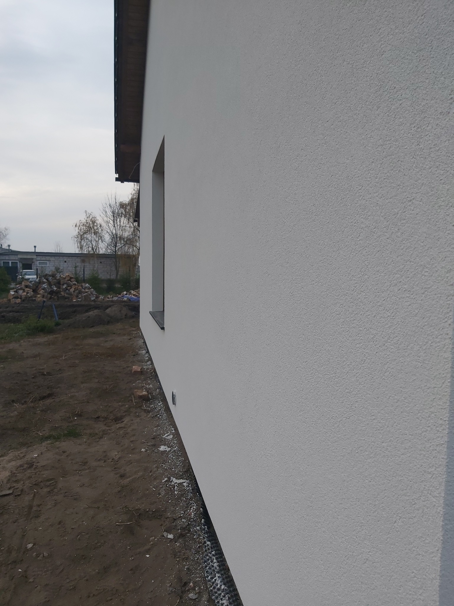 Boczny widok na świeżo otynkowaną, białą elewację domu jednorodzinnego z widocznym oknem i fragmentem dachu, teren wokół budynku jeszcze niezagospodarowany.