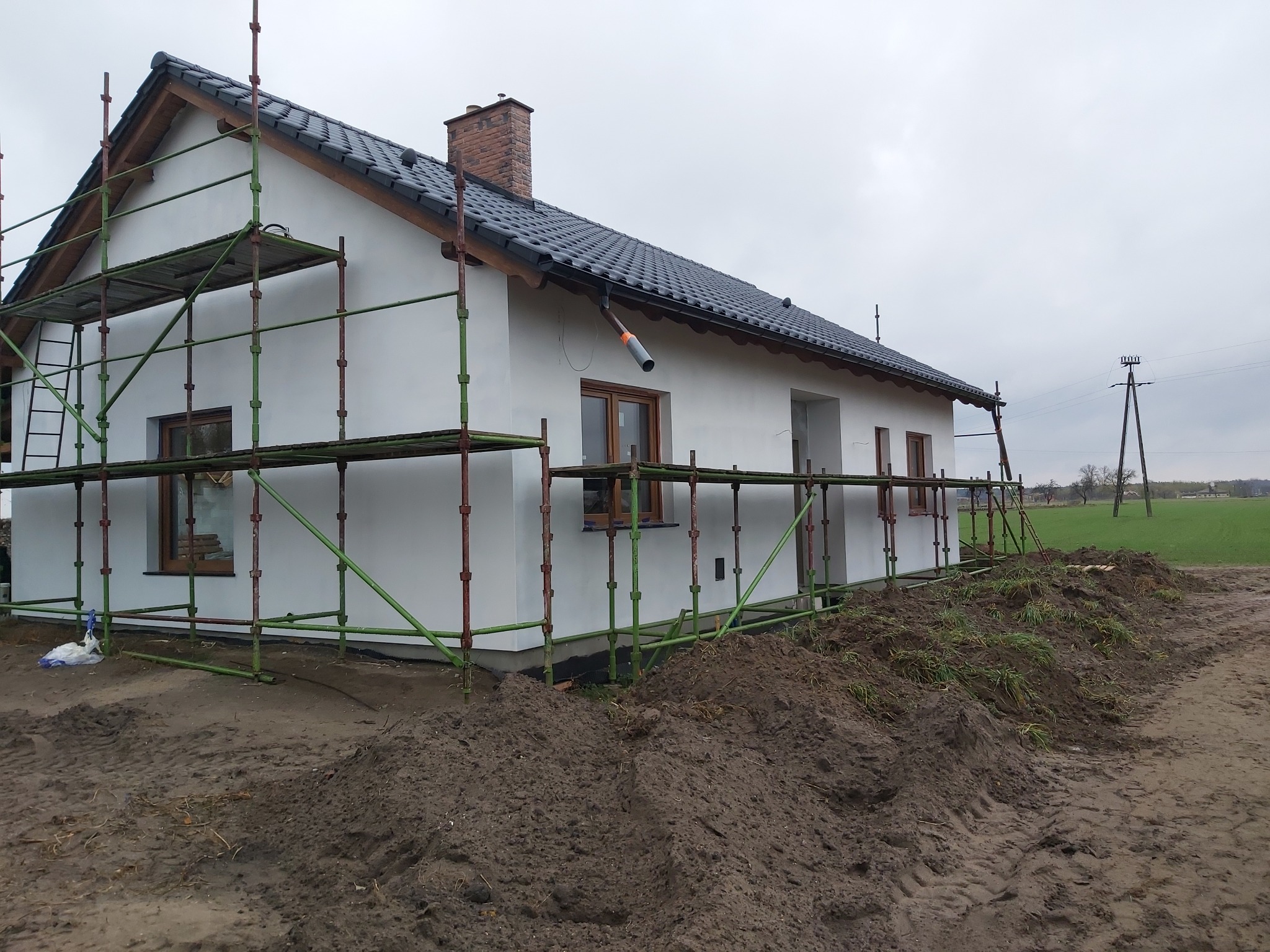 Dom jednorodzinny w trakcie prac elewacyjnych, z widocznymi rusztowaniami i świeżo otynkowaną fasadą w kolorze białym, na pierwszym planie hałda ziemi.