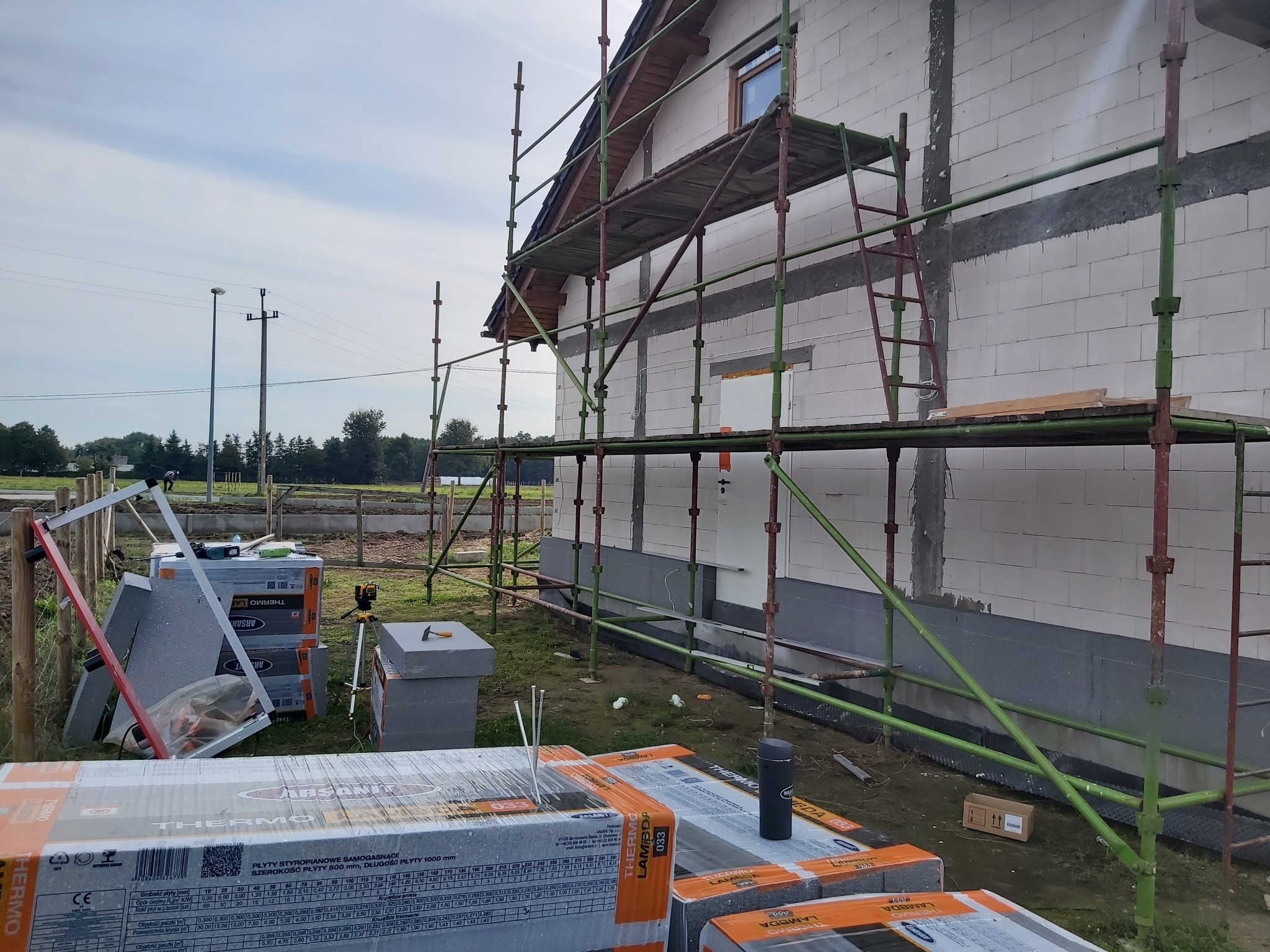 Budynek w trakcie ocieplania, widoczne rusztowanie, płyty styropianowe w oryginalnych opakowaniach, laserowy niwelator na statywie, ściany z betonu komórkowego częściowo ocieplone, szara podmurówka.