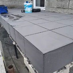 Szare płytki ułożone na balkonie w trakcie prac wykończeniowych, widoczne narzędzia i wiadro z farbą, fragment rusztowania.