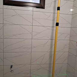 sowrem - Białe płytki imitujące marmur na ścianie łazienki, z widocznymi otworami na armaturę i poziomą linią lasera, żółta poziomica oparta o ścianę.