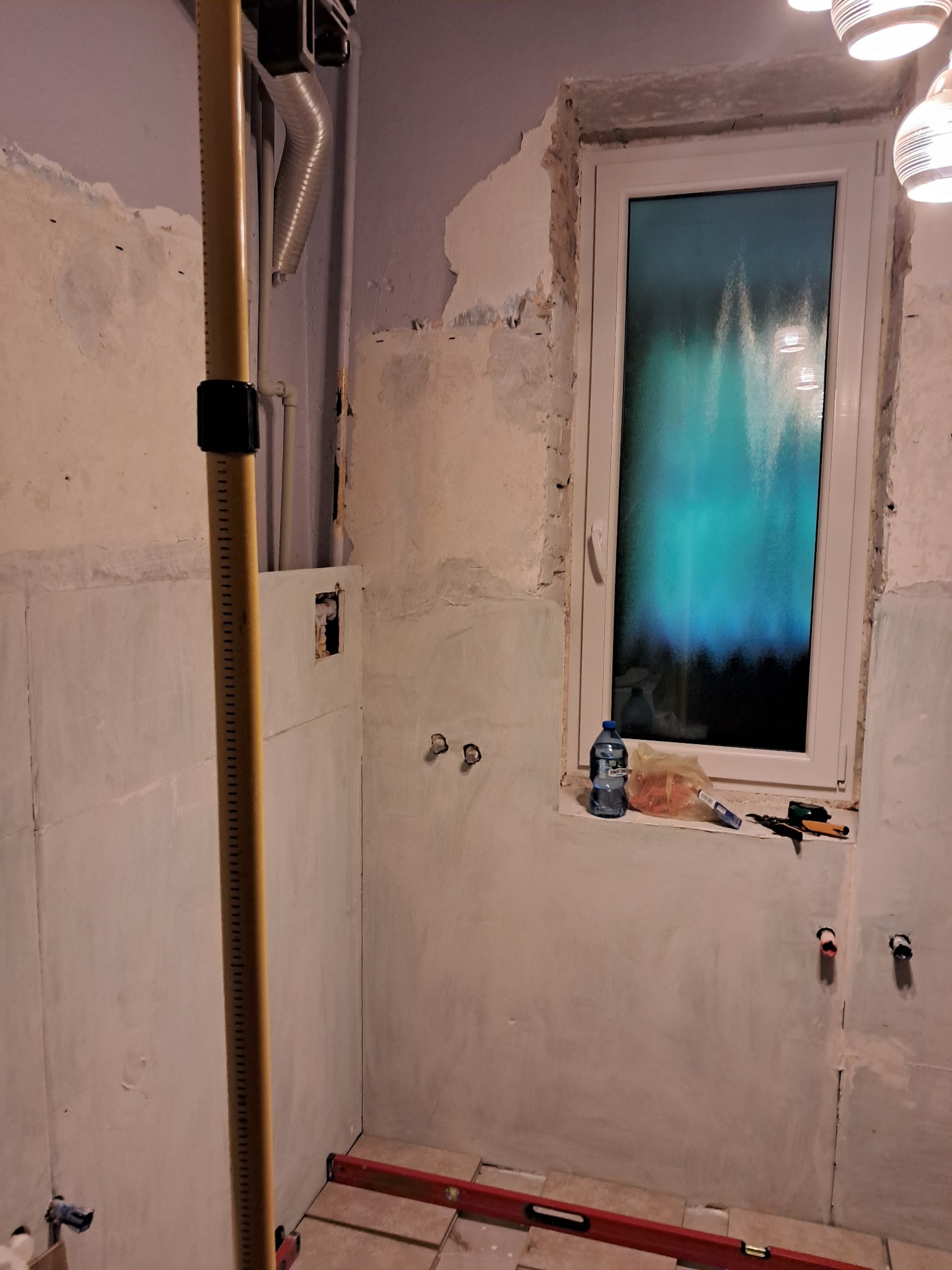 Łazienka w trakcie remontu w Myszkowie, widoczne przygotowane ściany, częściowo położone płytki podłogowe, narzędzia na parapecie i pionowa listwa pomiarowa.