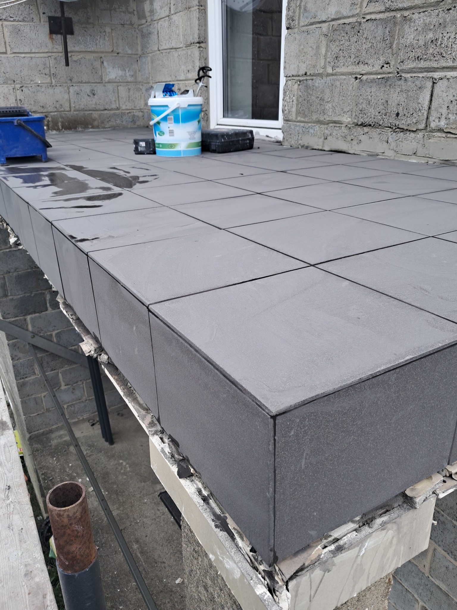 Szare płytki ułożone na balkonie w trakcie prac wykończeniowych, widoczne narzędzia i wiadro z farbą, fragment rusztowania.