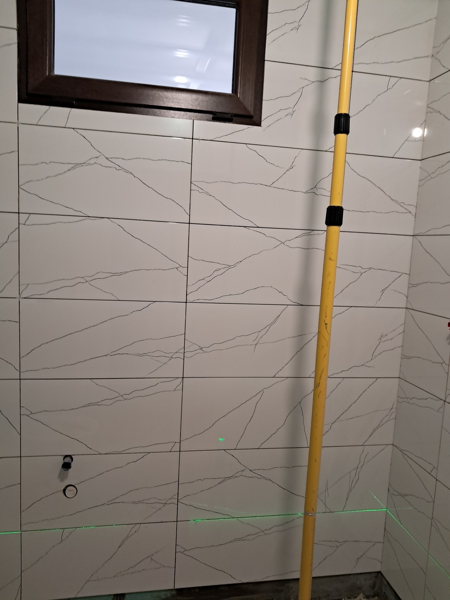 Białe płytki imitujące marmur na ścianie łazienki, z widocznymi otworami na armaturę i poziomą linią lasera, żółta poziomica oparta o ścianę.