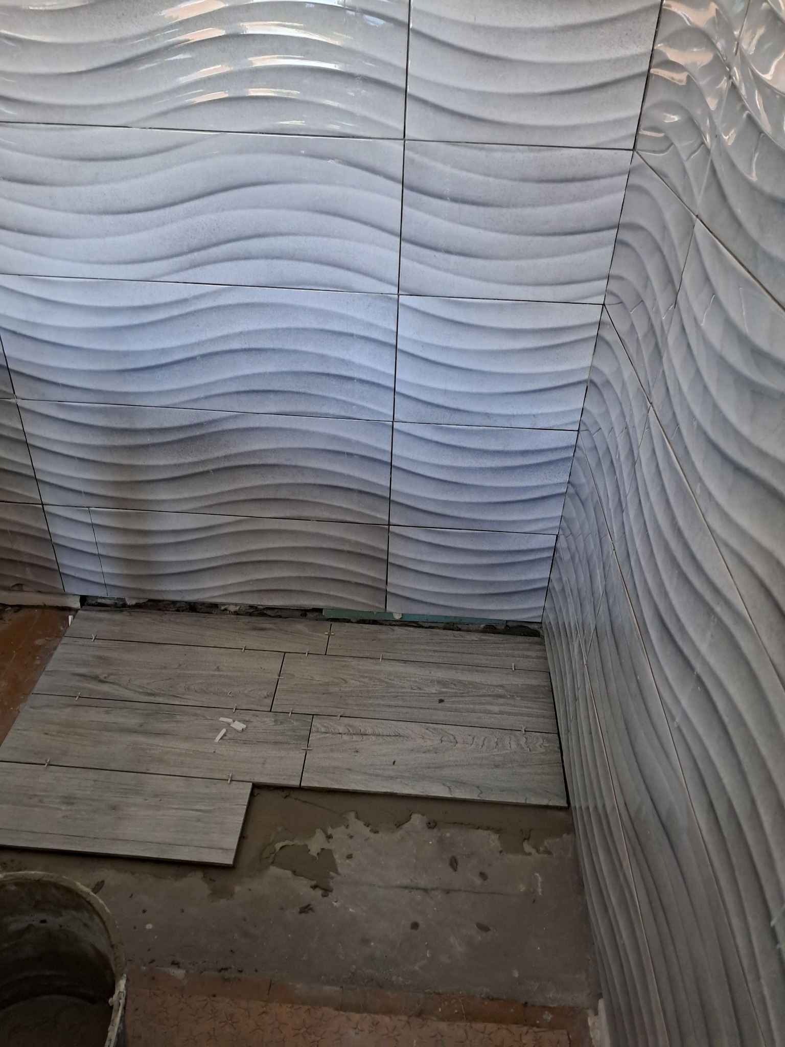 Układanie szarych płytek imitujących drewno na podłodze łazienki, z widocznymi płytkami ściennymi z falistym wzorem i zaprawą.