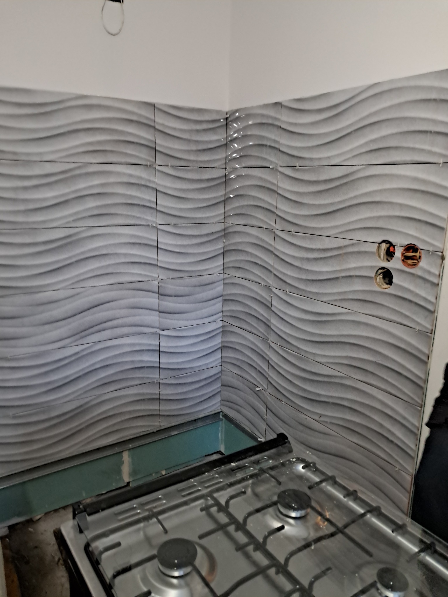 Ściana w kuchni wyłożona szarymi płytkami z falistym wzorem, widoczne otwory na gniazdka elektryczne i fragment kuchenki gazowej, etap prac wykończeniowych.