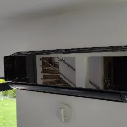 Czarny, błyszczący klimatyzator ścienny zamontowany w nowoczesnym wnętrzu, z widocznym odbiciem schodów i roślin w lustrzanej powierzchni urządzenia.