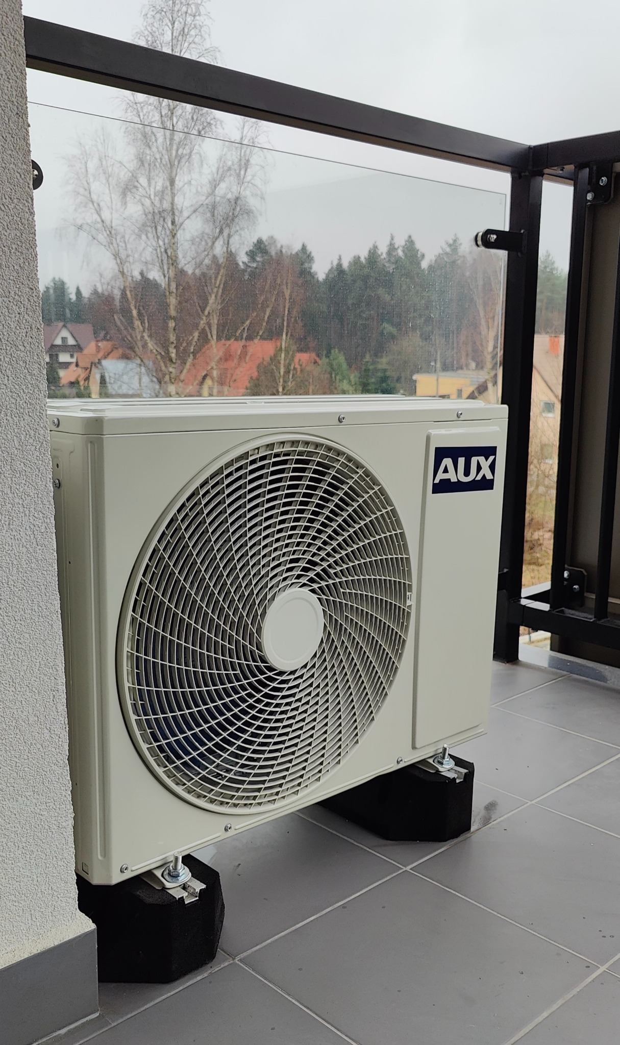 Zewnętrzna jednostka klimatyzacji marki AUX, zamontowana na balkonie z szarą podłogą i szklaną balustradą, widok na zewnątrz z drzewami i domami.