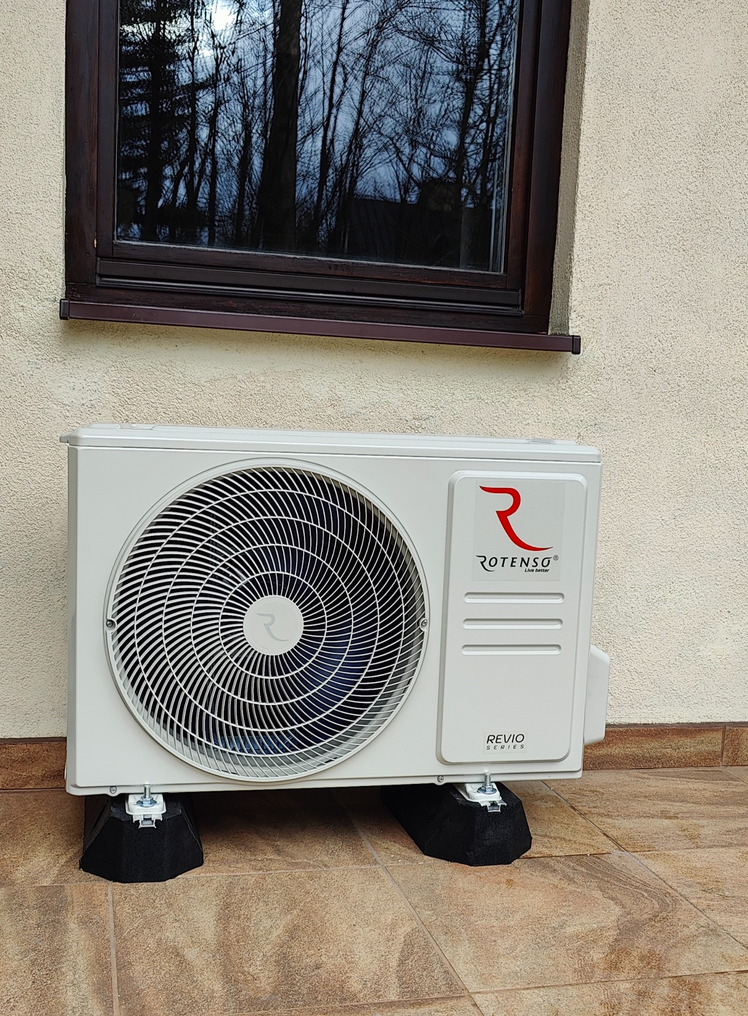 Zewnętrzna jednostka klimatyzacji marki Rotenso, model Revio, zamontowana na czarnych podkładkach antywibracyjnych pod oknem z odbiciem drzew.