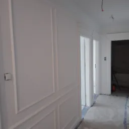 Korytarz w trakcie prac wykończeniowych: białe ściany z dekoracyjnymi panelami, widoczne kable wystające z sufitu, surowa podłoga zabezpieczona folią, otwarte drzwi prowadzące do kolejnych...