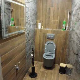 Szara wisząca toaleta w łazience wykończonej płytkami imitującymi drewno i marmur, z czarnym stojakiem na papier toaletowy i koszem na śmieci z bambusową pokrywką.