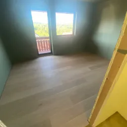 Pokój na poddaszu po remoncie, z jasnymi panelami podłogowymi, pomalowanymi na oliwkowo ścianami i widokiem na zieleń z balkonu przez okno.