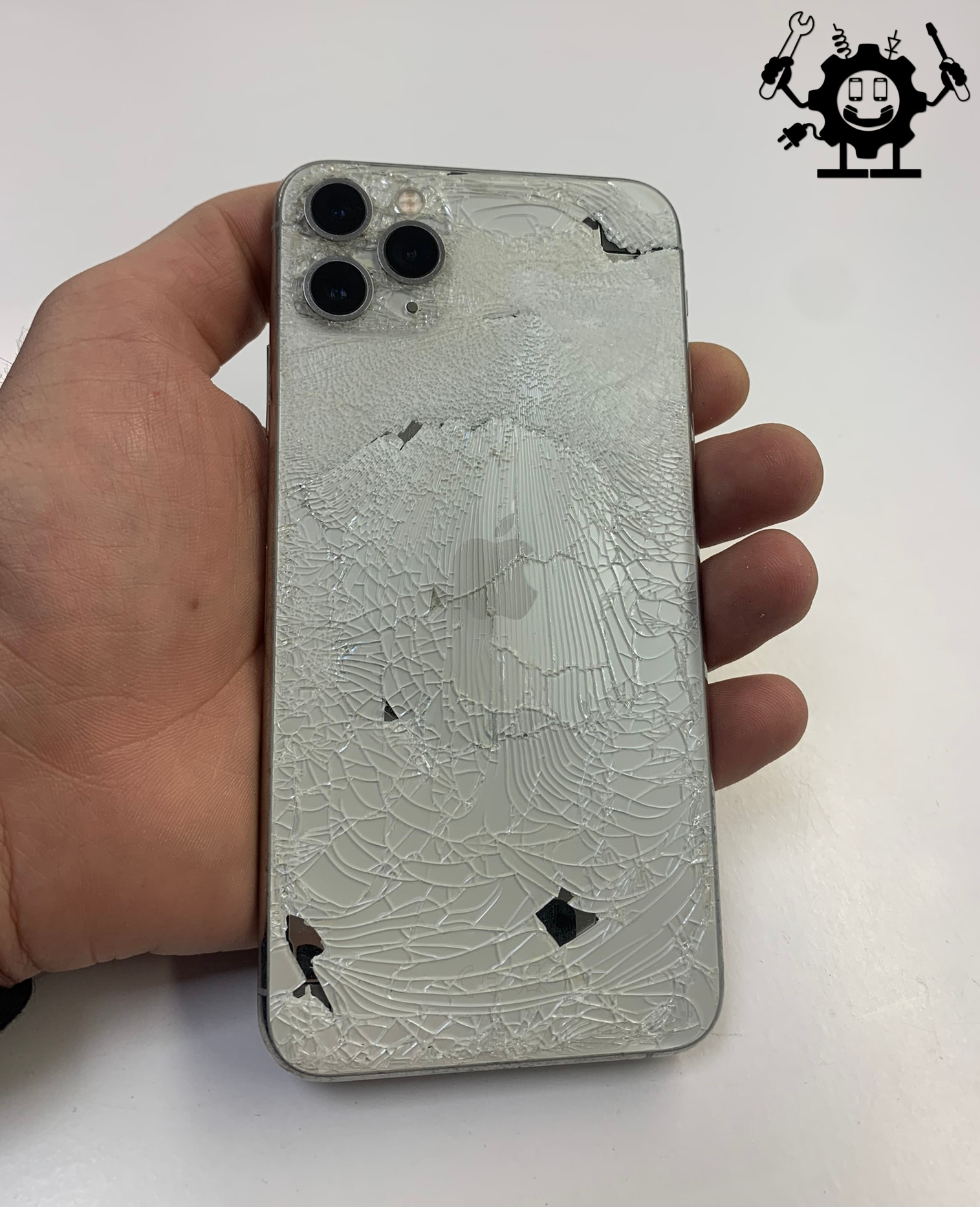 Dłoń trzyma iPhone 11 z bardzo mocno potłuczoną, srebrną obudową. Widoczne liczne pęknięcia i ubytki szkła. Logo Apple częściowo zniszczone.