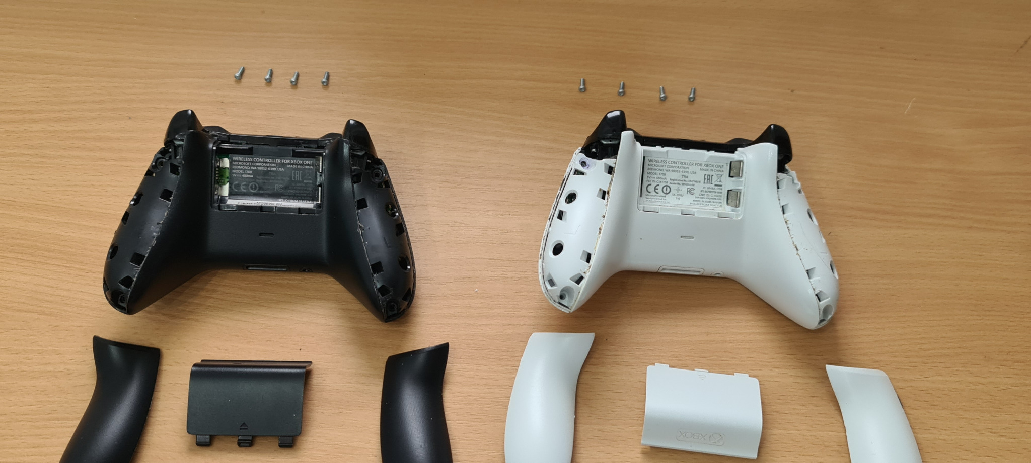 Dwa rozłożone kontrolery Xbox One: czarny i biały, z odkręconymi śrubkami u góry, leżą na drewnianym blacie.