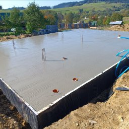 ARIS SPÓŁKA Z OGRANICZONĄ ODPOWIEDZIALNOŚCIĄ - Świeżo wylana płyta fundamentowa z wystającymi prętami zbrojeniowymi i widocznym drenażem, na tle pagórkowatego krajobrazu w słoneczny dzień.