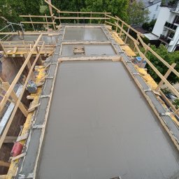 ARIS SPÓŁKA Z OGRANICZONĄ ODPOWIEDZIALNOŚCIĄ - Zalany beton na stropie budynku w trakcie budowy, widok z góry. Szalunki drewniane, częściowo widoczne budynki w tle. Świeży beton na całej powierzchni.