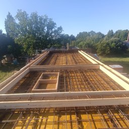 ARIS SPÓŁKA Z OGRANICZONĄ ODPOWIEDZIALNOŚCIĄ - Zbrojenie stropu z drewnianym szalunkiem, widok z góry na siatkę z prętów stalowych i deski, w tle zieleń drzew i błękitne niebo. Etap budowy domu.