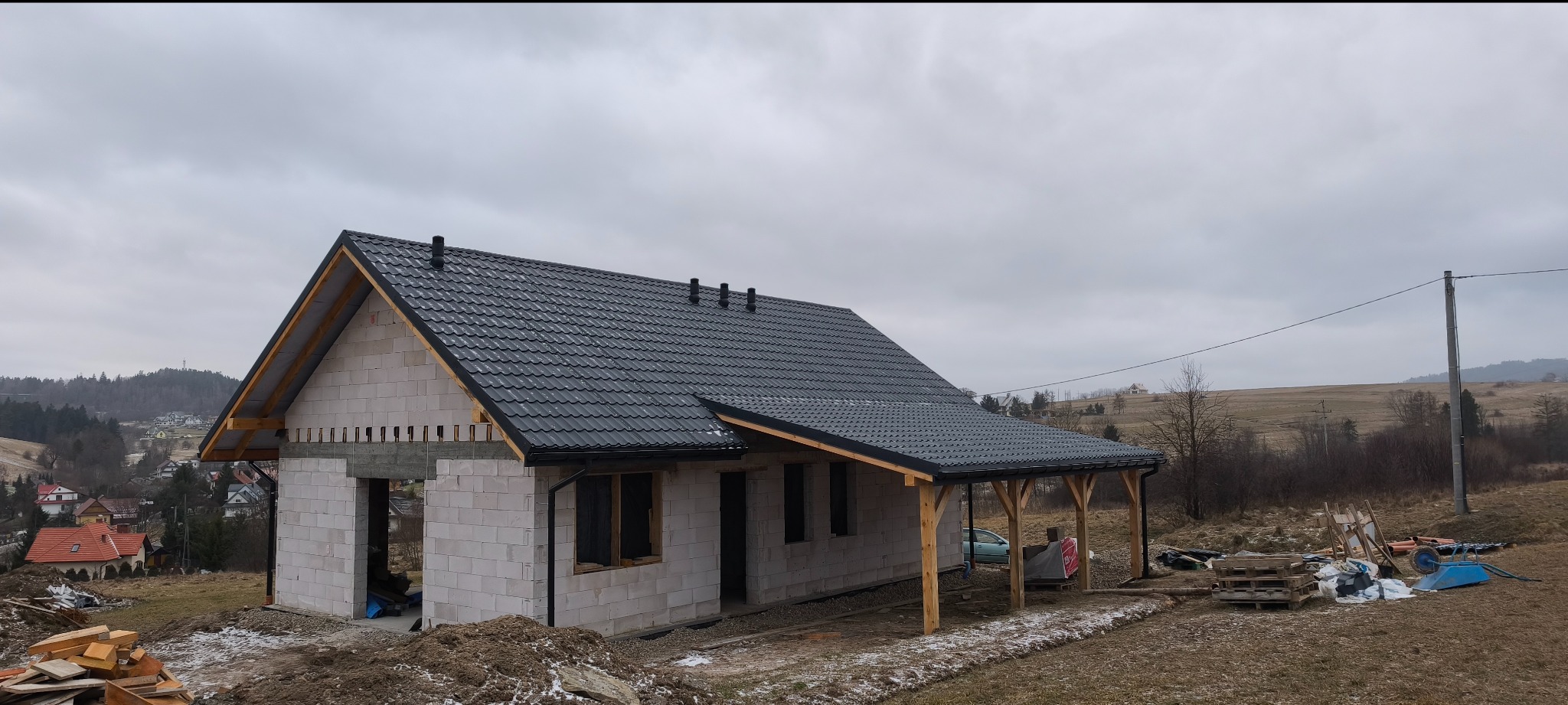 Niedokończony dom z białego bloczka z ciemnym dachem na tle pochmurnego nieba i pagórkowatego krajobrazu. Widoczna konstrukcja zadaszenia tarasu.
