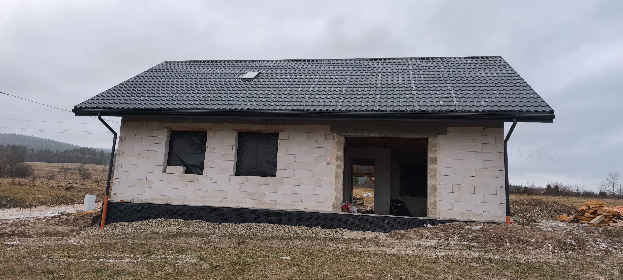 Niedokończony dom z białych bloczków, czarny dach z lukarną, orynnowanie. Okna zabite czarną folią. Widoczne fundamenty i teren budowy. Pochmurne niebo w tle.