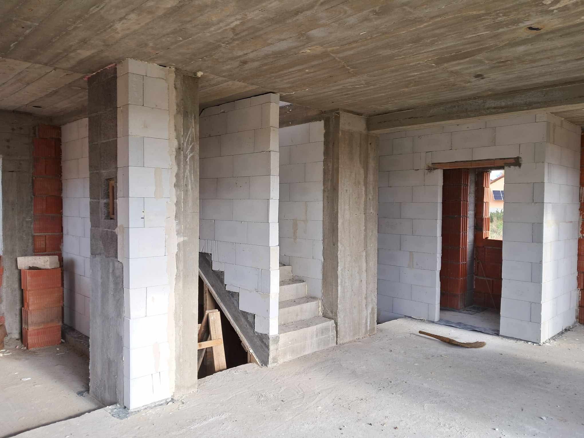 Wnętrze surowego domu w trakcie budowy: białe bloczki, betonowy strop, schody w stanie początkowym, widoczne elementy konstrukcyjne i narzędzie na podłodze.