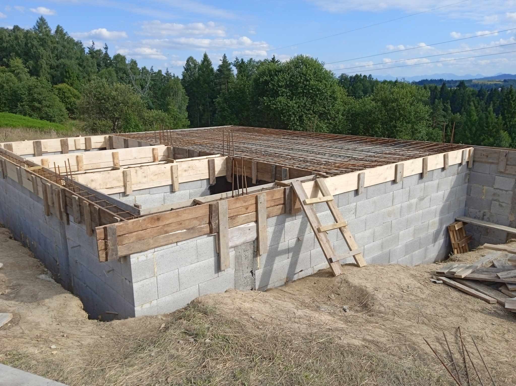 Fundamenty z bloczków betonowych, zbrojenie i drewniane szalunki w słoneczny dzień. W tle zalesione wzgórza i błękitne niebo. Widoczna drabina oparta o ścianę.