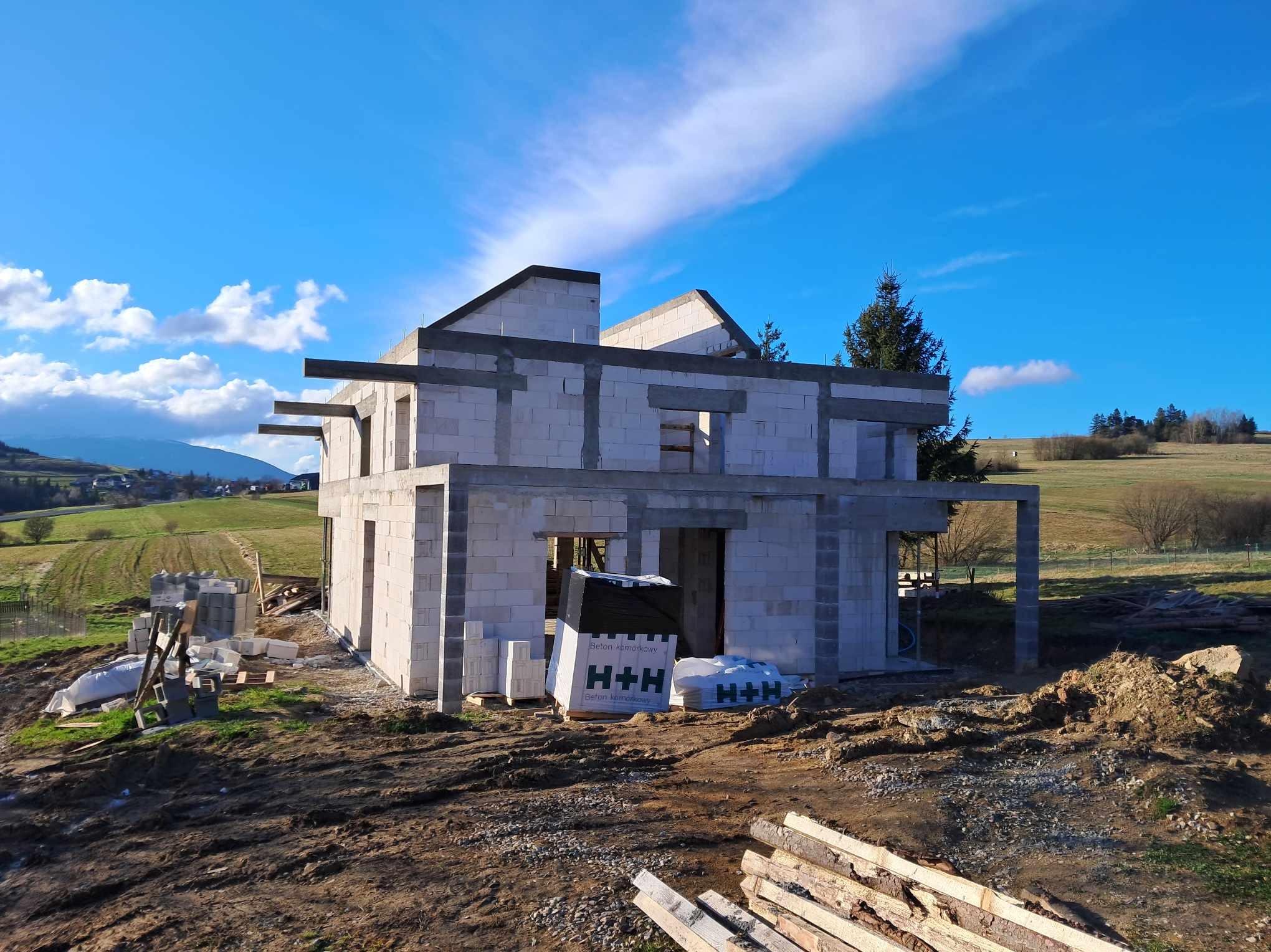 Surowy stan domu z betonu komórkowego H+H w malowniczej scenerii. Widoczne niezadaszone ściany, otwory okienne i drzwiowe. W tle pagórkowaty krajobraz pod błękitnym niebem.