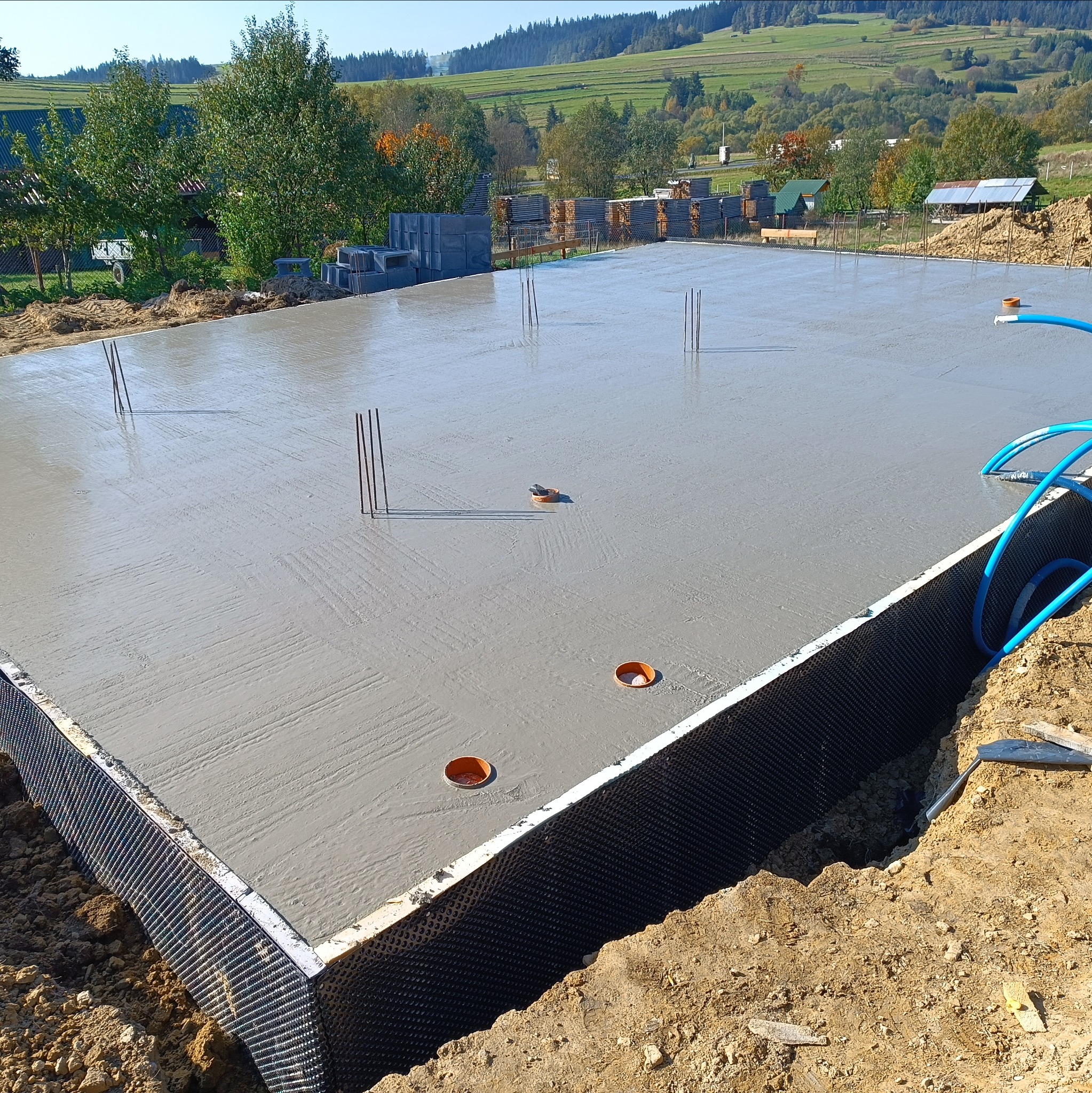 Świeżo wylana płyta fundamentowa z wystającymi prętami zbrojeniowymi i widocznym drenażem, na tle pagórkowatego krajobrazu w słoneczny dzień.