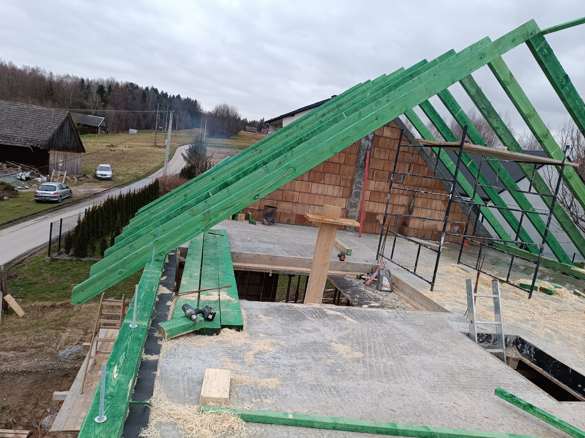Wznoszenie konstrukcji dachu z zielonych belek na budynku z czerwonej cegły. Widoczne narzędzia i elementy konstrukcyjne. W tle zabudowa wiejska i pochmurne niebo.