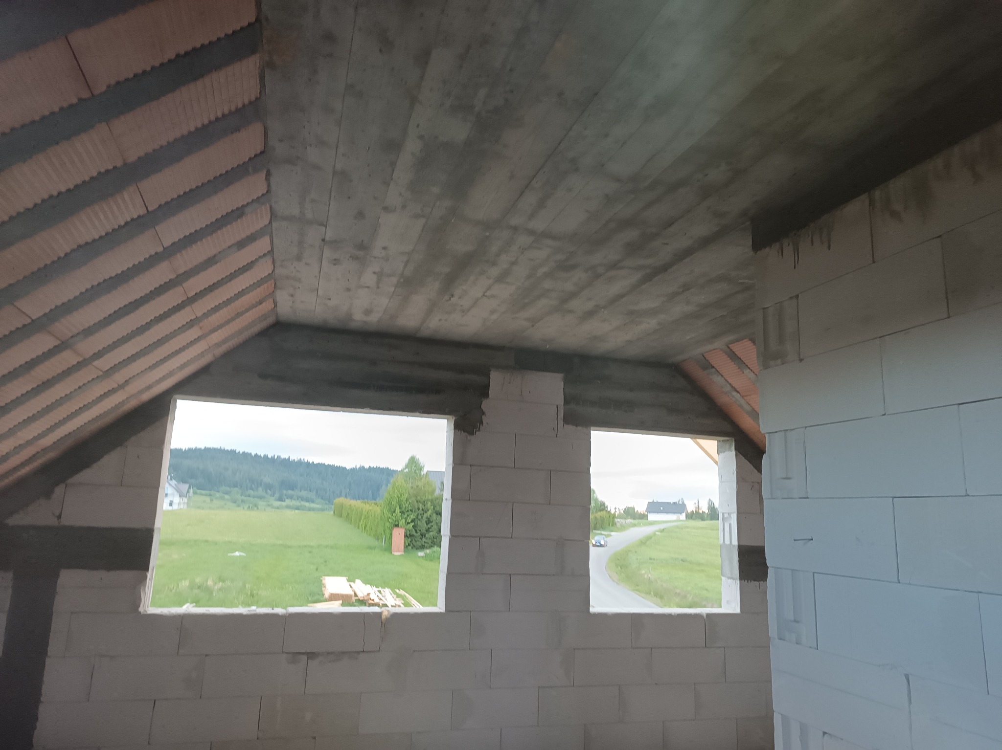 Surowy stan poddasza domu w budowie z widokiem na zewnątrz przez dwa otwory okienne. Widoczne bloczki, betonowy strop i fragment dachu. Krajobraz w tle.